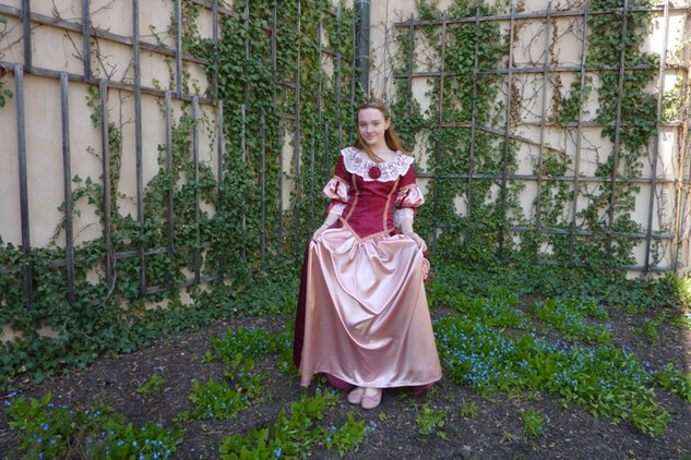 Princezna s korunkou v růžovo-červených šatech. Stojí v zámecké zahradě. | © Státní zámek Mníšek pod Brdy
