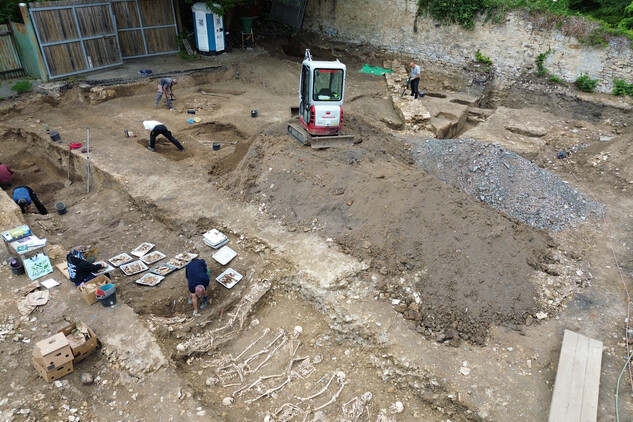 Finální vyzvednutí (exkavace) kosterních ostatků z liniového hrobu č. 12 a 17 a dvou skupinových u vjezdu na pozemek. Vlevo dole ukládání kosterních ostatků na podnosy a jejich popis antropoložkou (foto Egon Ditmar, 9. 6. 2025).